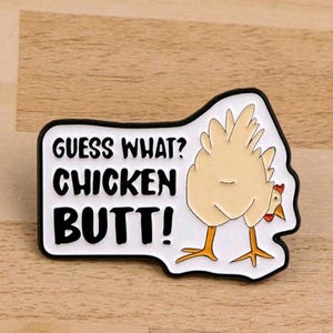 May include: A rectangular pin with a black border and the text "GUESS WHAT? CHICKEN BUTT!" in black letters on a white background. The pin features a cartoon chicken with its rear end facing forward.