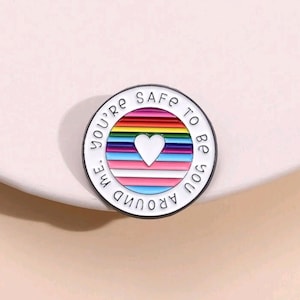 May include: White enamel pin with a rainbow and a heart in the center. The text around the pin reads "You're safe to be around me".