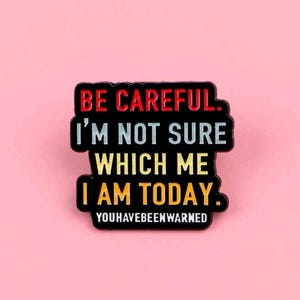 May include: A black pin with the text "BE CAREFUL. I'M NOT SURE WHICH ME I AM TODAY. YOUHAVEBEENWARNED" in red, white, and yellow. The pin is rectangular with rounded corners and is set against a pink background.
