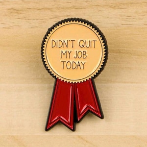 May include: A round, tan-colored pin with the text "DIDN'T QUIT MY JOB TODAY" in black lettering. The pin has a black border with a jagged edge and red ribbon streamers.