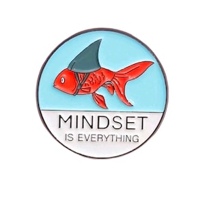 May include: A circular enamel pin featuring a bright orange goldfish wearing a dark gray shark fin on its back. The background is light blue, and the bottom section is white with the black text "MINDSET IS EVERYTHING" printed clearly.