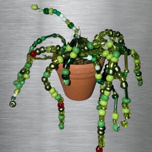 May include: A small terracotta pot containing a decorative plant sculpture made of numerous green, lime, and olive-coloured glass beads strung on flexible wire. The beaded strands drape over the sides of the pot, accented by small red beads.