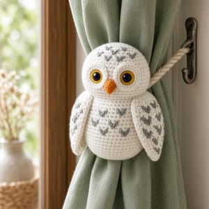 Puede incluir: Un alzapaños de cortina con forma de búho tejido en color blanco, con detalles de plumas en forma de V gris, grandes ojos negros y amarillos, y un pico naranja. El búho está unido a una cuerda trenzada que sujeta una cortina verde salvia.