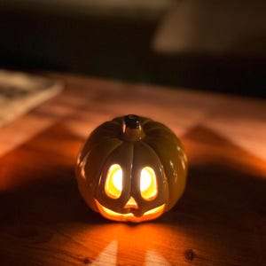 Könnte beinhalten: Eine gelbe Kürbislaterne aus Keramik mit einem geschnitzten Jack-o'-Lantern-Gesicht. Der Kürbis hat eine glatte, glänzende Oberfläche und einen dunklen Stiel an der Oberseite. Ein warmes, leuchtendes Licht scheint aus dem Inneren und beleuchtet die ausgeschnittenen Augen und den Mund.