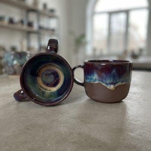 Op de afbeelding: Twee keramische mokken met een donkerbruine matte basis en een glanzende, druipende glazuurafwerking in tinten dieppaars, blauw en groen. De ene mok toont het spiraalpatroon aan de binnenkant, de andere mok toont het zijprofiel.