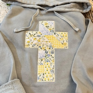 May include: A pale grey hoodie featuring a floral cross design. The cross is made from fabric patches with yellow gingham and floral patterns. The hoodie has a drawstring hood and cuffed sleeves.