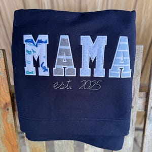 May include: A navy blue sweatshirt featuring the word MAMA in large, appliqued letters. The letters have varied patterns including sharks, grey stripes, light blue dots, and light blue stripes. Below the word, the text est. 2025 is embroidered.