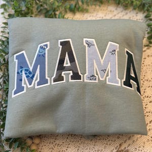 May include: A sage green sweatshirt featuring the word MAMA in large, arched, collegiate-style letters. Each letter has a unique pattern: the first M is blue with turtles, the A is camouflage, the second M is light blue with dinosaurs, and the final A is dark green.