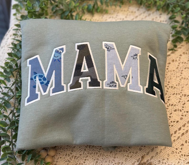May include: A sage green sweatshirt featuring the word MAMA in large, arched, collegiate-style letters. Each letter has a unique pattern: the first M is blue with turtles, the A is camouflage, the second M is light blue with dinosaurs, and the final A is dark green.