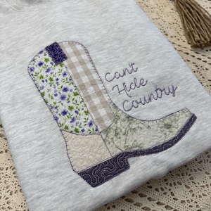 Puede incluir: Una sudadera gris claro con un diseño de patchwork de una bota de vaquero. La bota incorpora telas florales, de cuadros y estampadas en tonos morados, verdes y beige. El texto "Can't Hide Country" está bordado en morado.