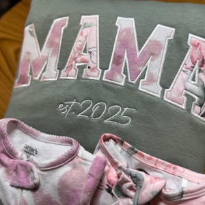 May include: An olive green sweatshirt featuring the word MAMA in large block letters with a pink floral pattern, and the text est. 2025 embroidered below. Two white baby sleepers with a pink floral print are positioned in front of the sweatshirt.