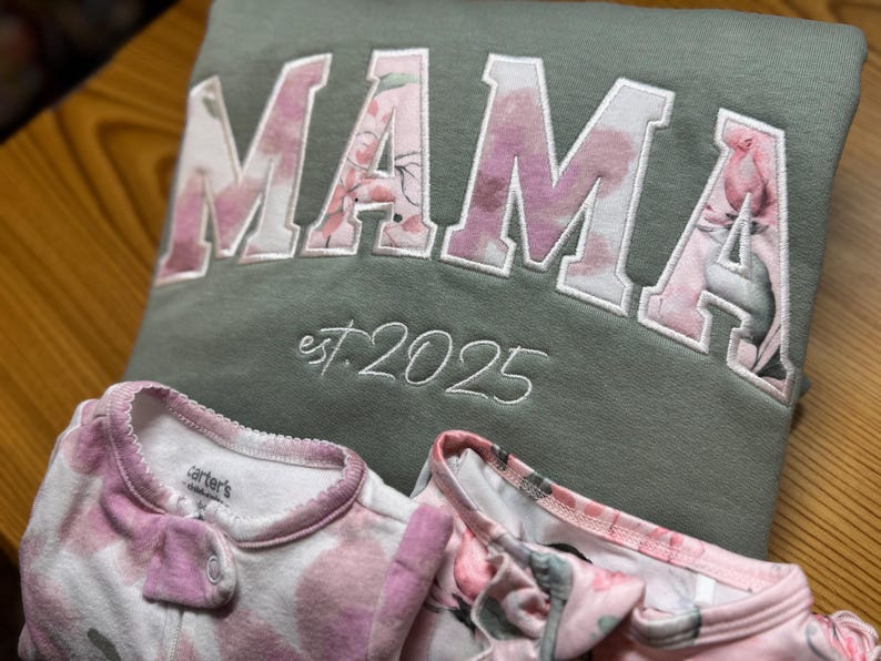 May include: An olive green sweatshirt featuring the word MAMA in large block letters with a pink floral pattern, and the text est. 2025 embroidered below. Two white baby sleepers with a pink floral print are positioned in front of the sweatshirt.