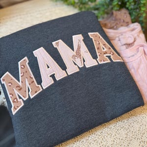 May include: A dark charcoal gray sweatshirt featuring the word MAMA in large, block-style letters. The letters are appliqued with a mix of light pink fabric and a brown leopard print pattern, all outlined with a white stitched border.