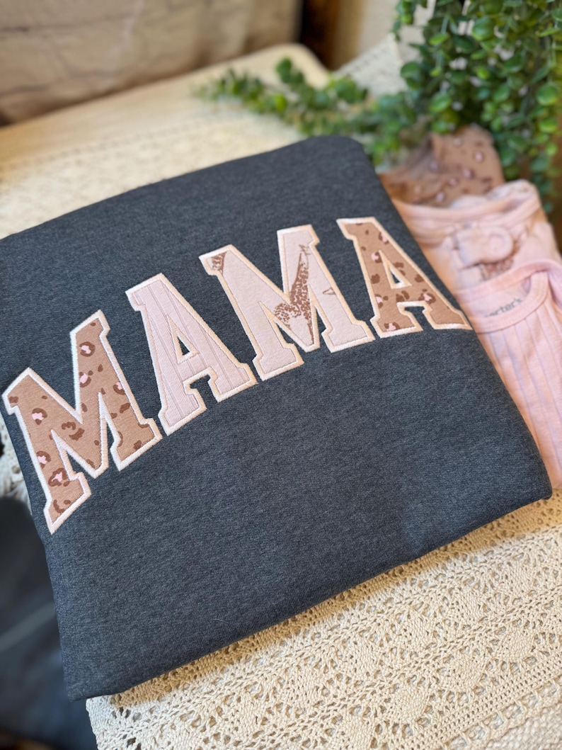 May include: A dark charcoal gray sweatshirt featuring the word MAMA in large, block-style letters. The letters are appliqued with a mix of light pink fabric and a brown leopard print pattern, all outlined with a white stitched border.