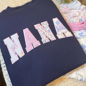 May include: A navy blue sweatshirt featuring the word NANA in large block letters. Each letter is filled with a light-colored fabric pattern of small flowers and butterflies, outlined with a thin pink border, creating a distinct contrast.