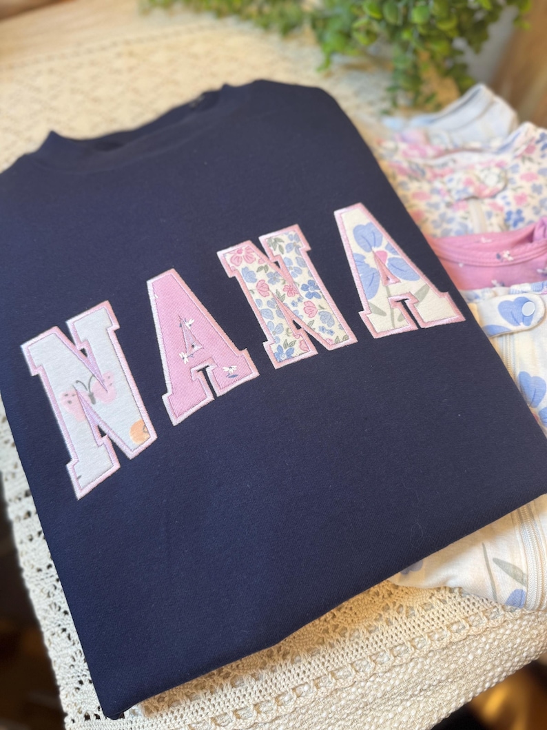 May include: A navy blue sweatshirt featuring the word NANA in large block letters. Each letter is filled with a light-colored fabric pattern of small flowers and butterflies, outlined with a thin pink border, creating a distinct contrast.