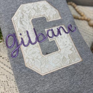 Pode incluir: Uma camisola cinzenta mesclada com uma letra G grande em estilo bloco aplicada em renda floral branca. O nome "gilbane" está bordado no centro da letra numa escrita cursiva roxa com um contorno de fio roxo escuro.
