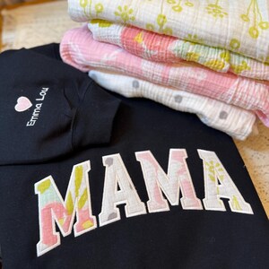 May include: A black sweatshirt featuring the word MAMA in large block letters with a floral pattern fill, and a sleeve embroidered with a pink heart and the text Emma Lou. Beside it are three folded muslin blankets with yellow and pink floral prints.