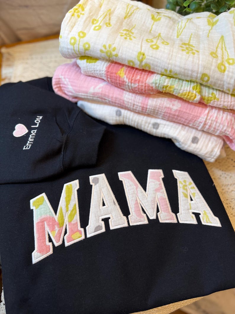 May include: A black sweatshirt featuring the word MAMA in large block letters with a floral pattern fill, and a sleeve embroidered with a pink heart and the text Emma Lou. Beside it are three folded muslin blankets with yellow and pink floral prints.