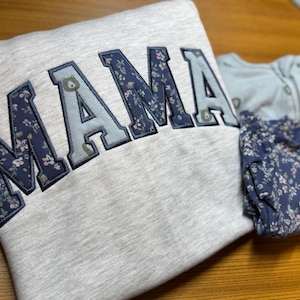 May include: A light gray sweatshirt featuring the word MAMA in large, varsity-style applique letters with alternating dark blue floral and light blue bear-patterned fabric, alongside a matching dark blue floral patterned infant sleeper garment.