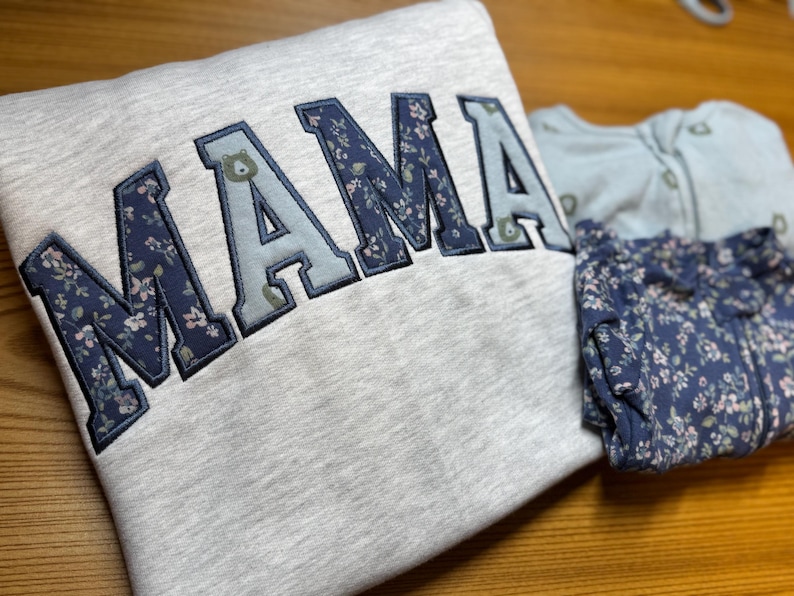 May include: A light gray sweatshirt featuring the word MAMA in large, varsity-style applique letters with alternating dark blue floral and light blue bear-patterned fabric, alongside a matching dark blue floral patterned infant sleeper garment.