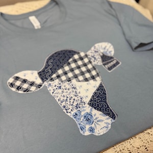 Può includere: T-shirt azzurra con un design a testa di mucca patchwork. La testa di mucca è composta da vari tessuti a fantasia blu e bianca, tra cui quadretti, floreali e paisley. La maglietta ha un girocollo e maniche corte.