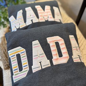 May include: Two dark charcoal gray sweatshirts featuring large, block-lettered appliqu&eacute;s. The top sweatshirt displays the word MAMA, and the bottom sweatshirt displays the word DADA. The letters feature various pastel-colored horizontal stripes.
