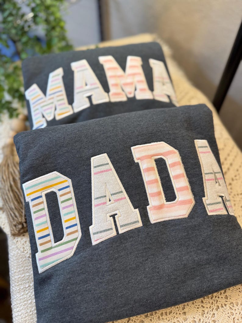 May include: Two dark charcoal gray sweatshirts featuring large, block-lettered appliqu&eacute;s. The top sweatshirt displays the word MAMA, and the bottom sweatshirt displays the word DADA. The letters feature various pastel-colored horizontal stripes.