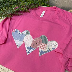 Puede incluir: Una camiseta rosa brillante con cuatro aplicaciones en forma de corazón. Cada corazón está hecho de una tela estampada diferente, incluyendo diseños florales, cuadros y abejas. La camiseta tiene mangas cortas y cuello redondo.