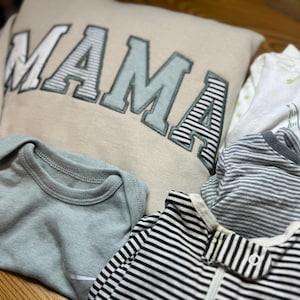 May include: A beige sweatshirt with the word MAMA in large block letters, a light green baby bodysuit, a white baby sleeper with green patterns, and two baby sleepers with black and white horizontal stripes and a zipper closure.