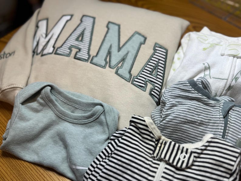 May include: A beige sweatshirt with the word MAMA in large block letters, a light green baby bodysuit, a white baby sleeper with green patterns, and two baby sleepers with black and white horizontal stripes and a zipper closure.