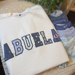 May include: A white crewneck sweatshirt featuring the word ABUELA in large, block-style letters. Each letter is outlined in dark blue and filled with a patterned fabric displaying various floral designs in shades of blue, yellow, and pink.