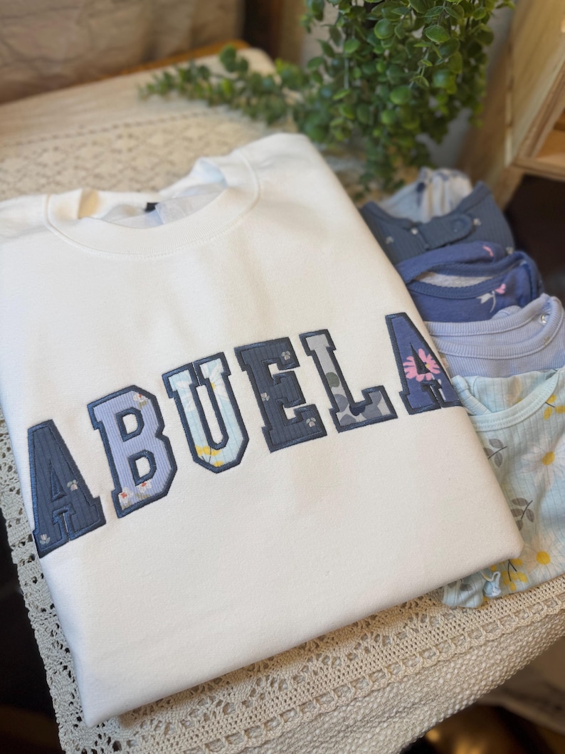 May include: A white crewneck sweatshirt featuring the word ABUELA in large, block-style letters. Each letter is outlined in dark blue and filled with a patterned fabric displaying various floral designs in shades of blue, yellow, and pink.