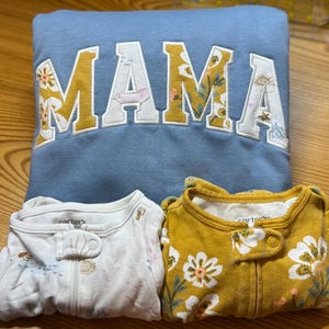 May include: A light blue sweatshirt with the word MAMA appliqued in white and mustard yellow floral fabric. Below are two infant sleepers: one white with small sea creature prints and one mustard yellow with a white floral pattern by Carter's.