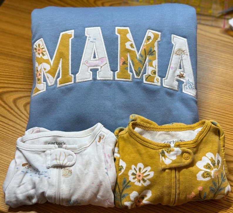 May include: A light blue sweatshirt with the word MAMA appliqued in white and mustard yellow floral fabric. Below are two infant sleepers: one white with small sea creature prints and one mustard yellow with a white floral pattern by Carter's.