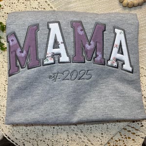 May include: A heather gray sweatshirt featuring the word MAMA in large, block-style letters. The letters alternate between a solid purple and a white floral pattern. Below the word, the text est. 2025 is embroidered in a dark gray script font.