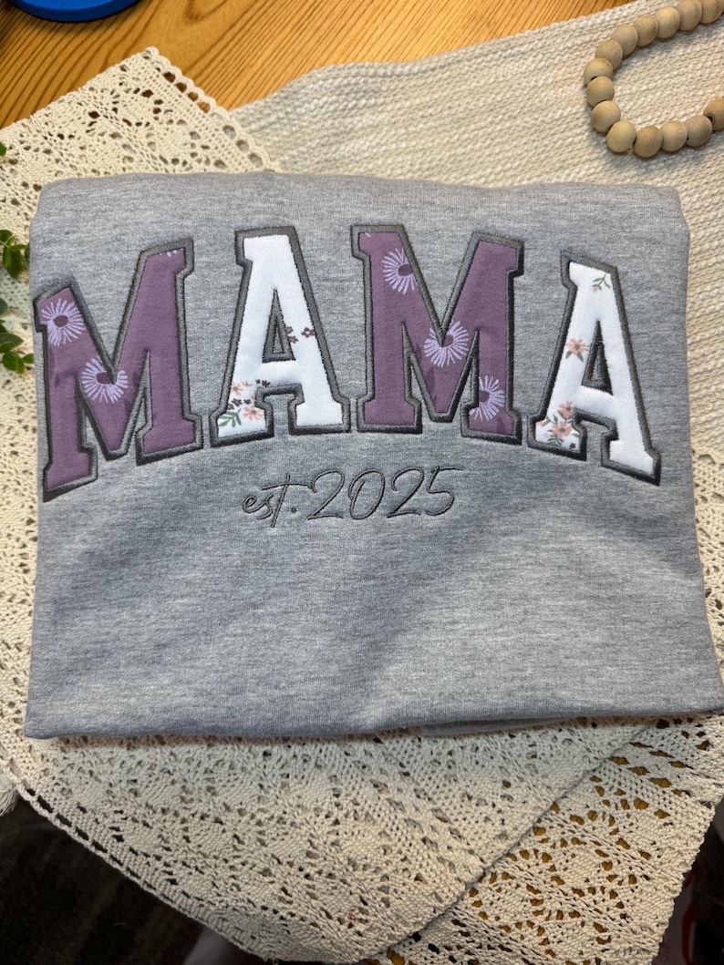 May include: A heather gray sweatshirt featuring the word MAMA in large, block-style letters. The letters alternate between a solid purple and a white floral pattern. Below the word, the text est. 2025 is embroidered in a dark gray script font.