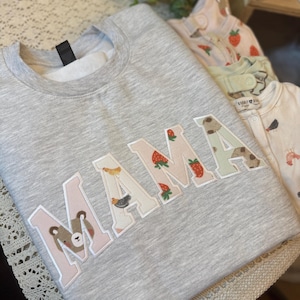 May include: A heather gray crewneck sweatshirt featuring the word MAMA in large block letters. Each letter is filled with a different pattern, including a bear face, chickens, strawberries, and a light green print, all outlined in white stitching.