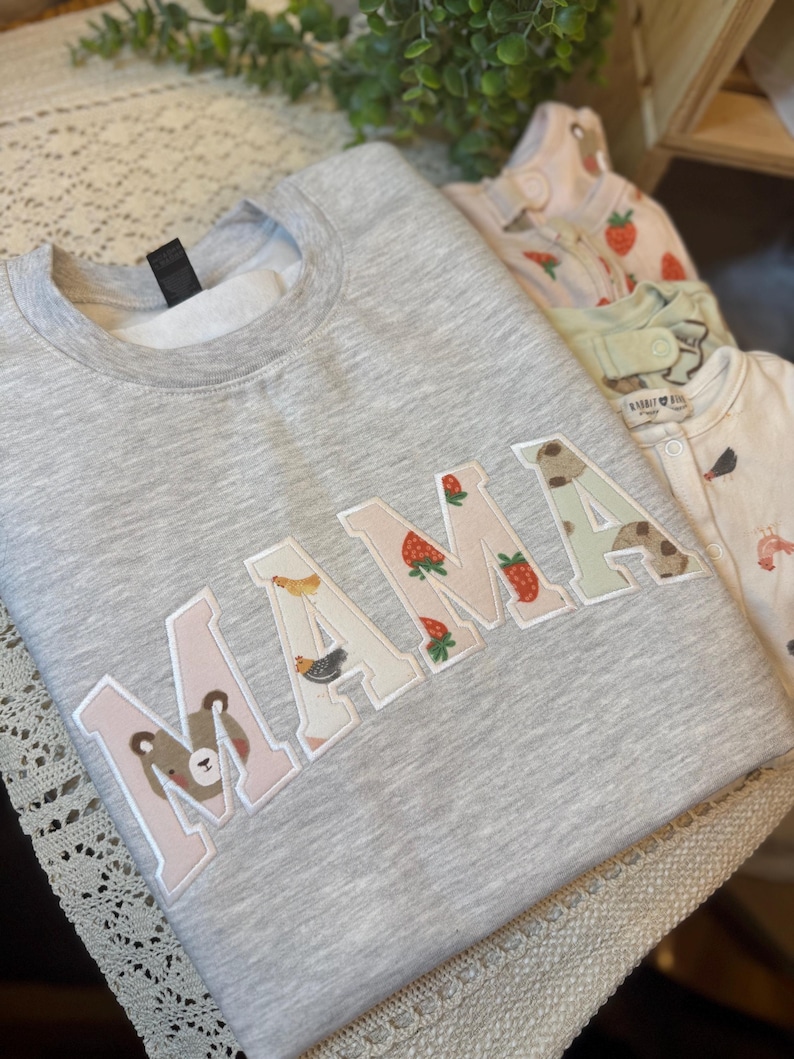 May include: A heather gray crewneck sweatshirt featuring the word MAMA in large block letters. Each letter is filled with a different pattern, including a bear face, chickens, strawberries, and a light green print, all outlined in white stitching.
