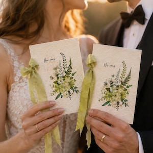 May include: Two rectangular vow booklets made of textured off-white paper, each featuring a botanical illustration of green ferns and flowers. The left booklet is labeled "Her vows" and the right "His vows," both tied with frayed light green silk ribbons.