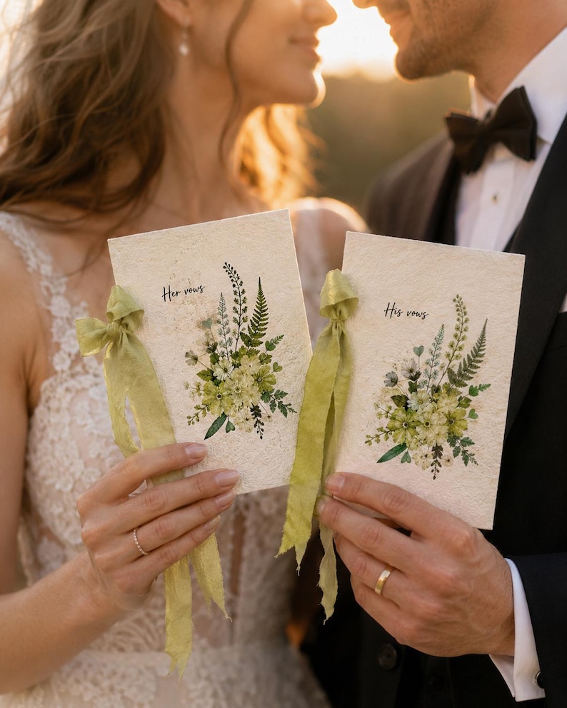 May include: Two rectangular vow booklets made of textured off-white paper, each featuring a botanical illustration of green ferns and flowers. The left booklet is labeled "Her vows" and the right "His vows," both tied with frayed light green silk ribbons.