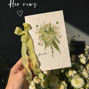 May include: A rectangular off-white textured paper card featuring a pressed green and white floral arrangement with a fern leaf. The card is tied with a light green ribbon and displays the text "Her vows" in a small black script font.