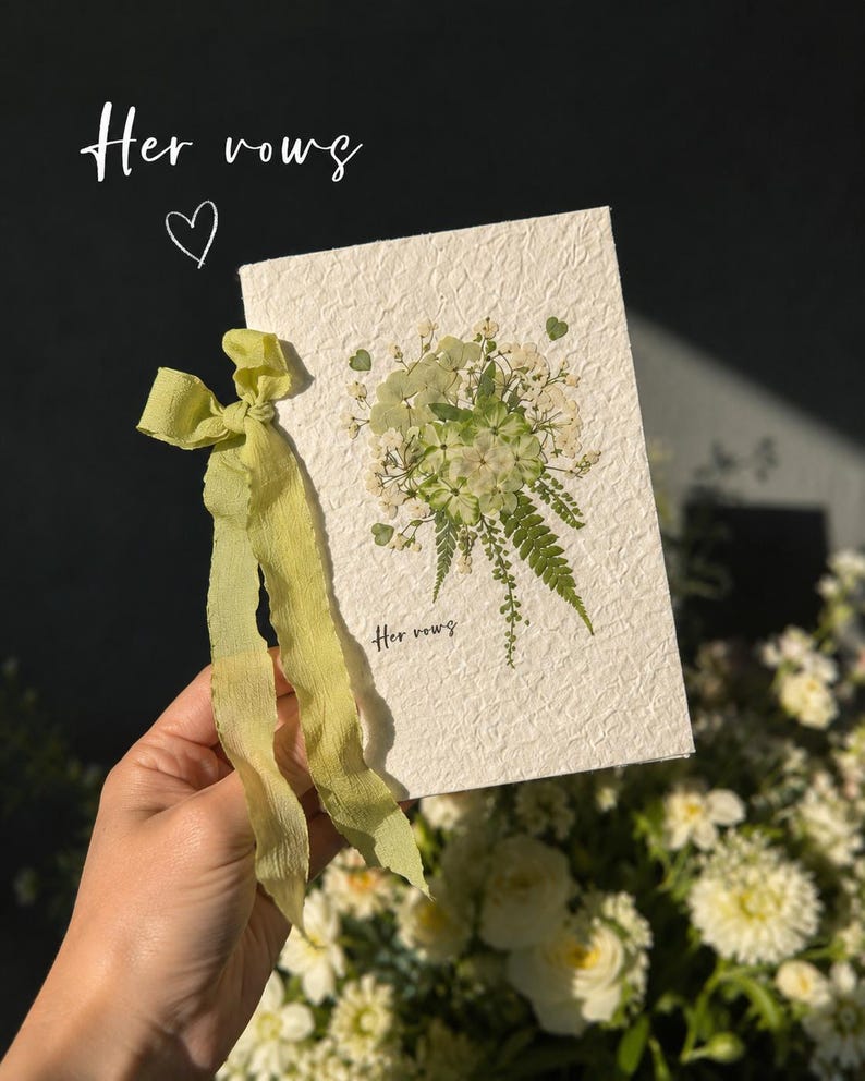 May include: A rectangular off-white textured paper card featuring a pressed green and white floral arrangement with a fern leaf. The card is tied with a light green ribbon and displays the text "Her vows" in a small black script font.
