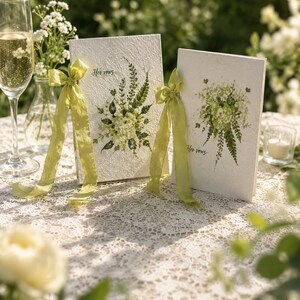May include: Two white textured vow books featuring a printed green botanical bouquet illustration and the text "Her vows" in black script. Each book is adorned with a light green silk ribbon tied in a bow on the left side of the cover.