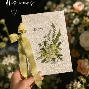 May include: A rectangular off-white textured paper booklet titled His vows, featuring a pressed green floral arrangement on the cover and a sage green ribbon tied to the side. The text His vows is printed in a small black script font on the front cover.