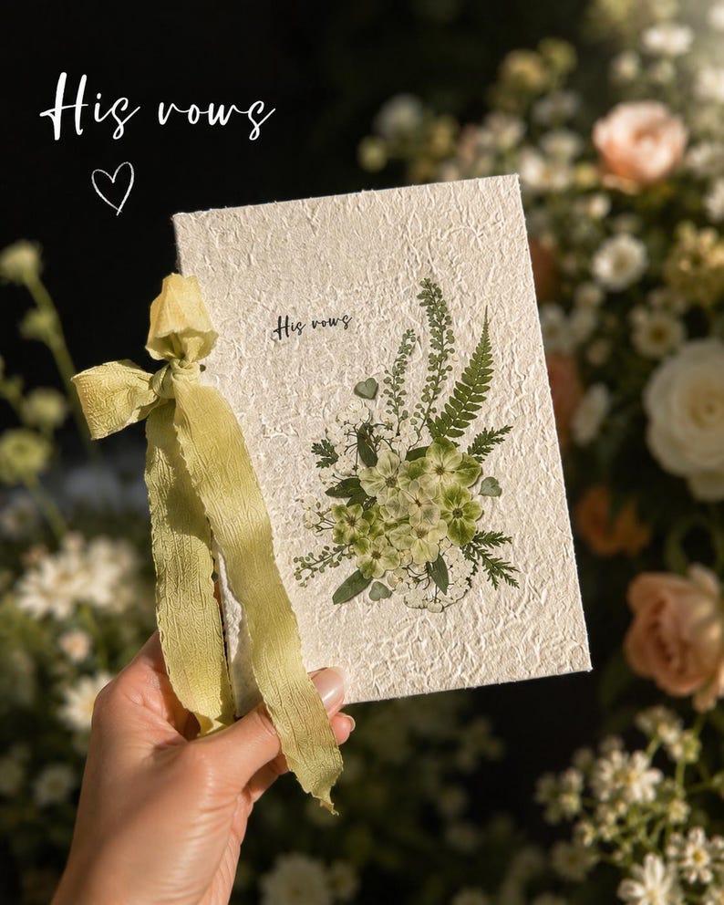 May include: A rectangular off-white textured paper booklet titled His vows, featuring a pressed green floral arrangement on the cover and a sage green ribbon tied to the side. The text His vows is printed in a small black script font on the front cover.
