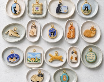 Może przedstawiać: Kolekcja siedemnastu małych, ręcznie malowanych ceramicznych zawieszek z motywem kota, z których każda przedstawia unikalne ilustracje kotów, motywy kwiatowe lub tekst, umieszczonych na pojedynczych, małych, kremowych talerzykach.