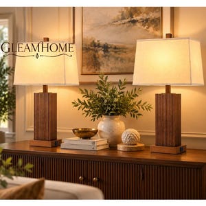 May include: Two table lamps with rectangular white fabric shades and vertical ribbed wooden bases. The lamps sit on a wooden sideboard, flanking a white ceramic vase filled with greenery and a small decorative sphere on stacked books.