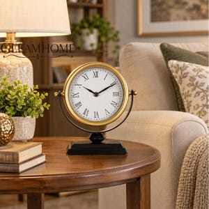 May include: A round table clock with a gold-toned frame and a white face featuring black Roman numerals. The clock is mounted on a black metal swivel stand with a rectangular base. The clock hands are black and thin, indicating the current time.