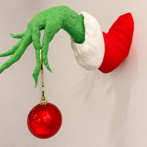 Puede incluir: Una escultura decorativa de pared que muestra una mano verde texturizada que emerge de una manga roja con un puño de piel blanca. Los dedos largos y delgados de la mano verde sostienen un adorno navideño rojo, redondo y brillante, con un hilo dorado.